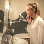 Side view of blond woman in white blouse standing in bathroom looking closely at face skin in small mirror