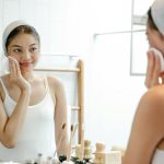 Smiling young Asian female looking at reflection in mirror while cleaning face skin with cotton pad during daily morning routine in bathroom